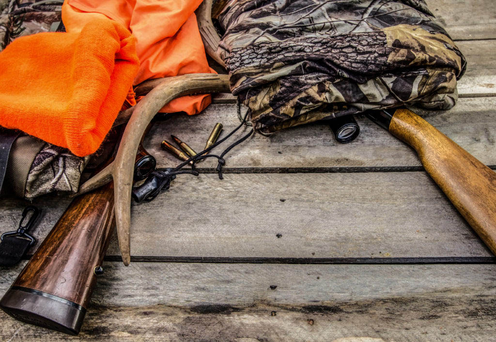 a pile of hunting gear including a rifle, blaze orange, bullets and more lying on a deck